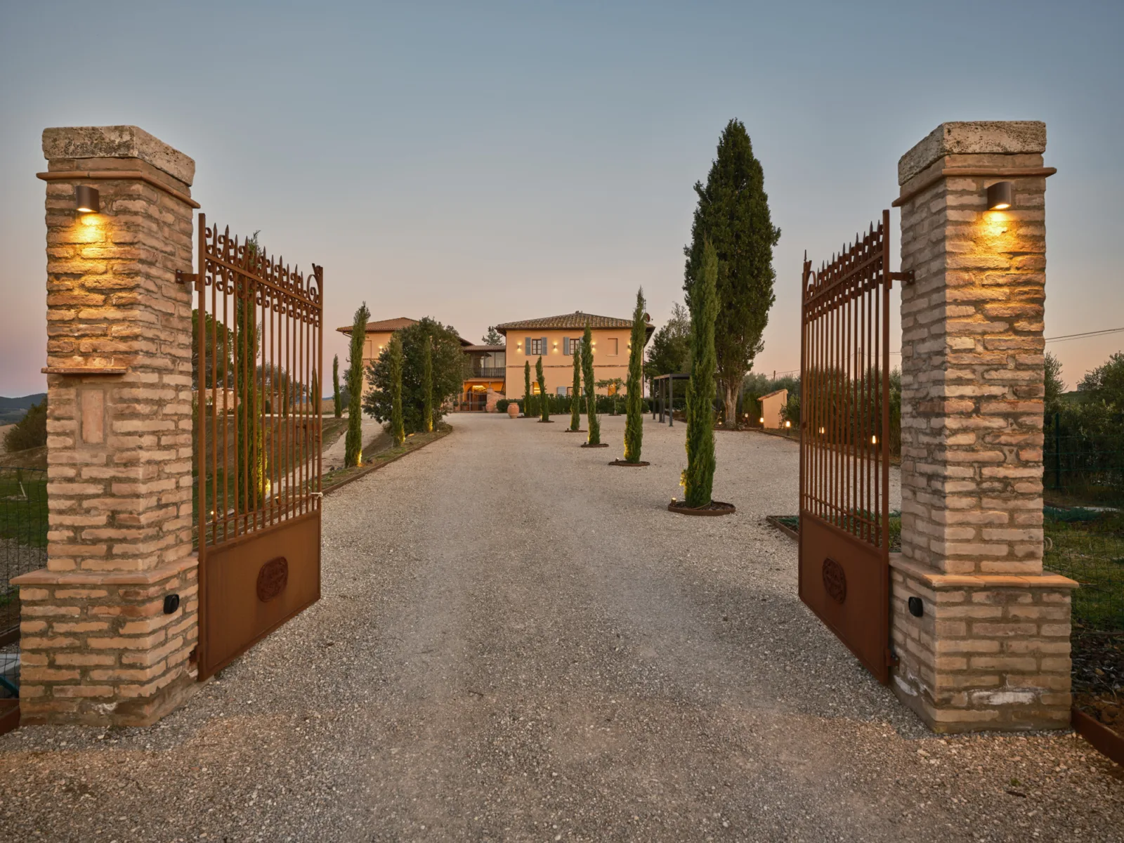 The gated approach to Villa Eos at twilight, lined with cypresses, with the restored farmhouse in the background.