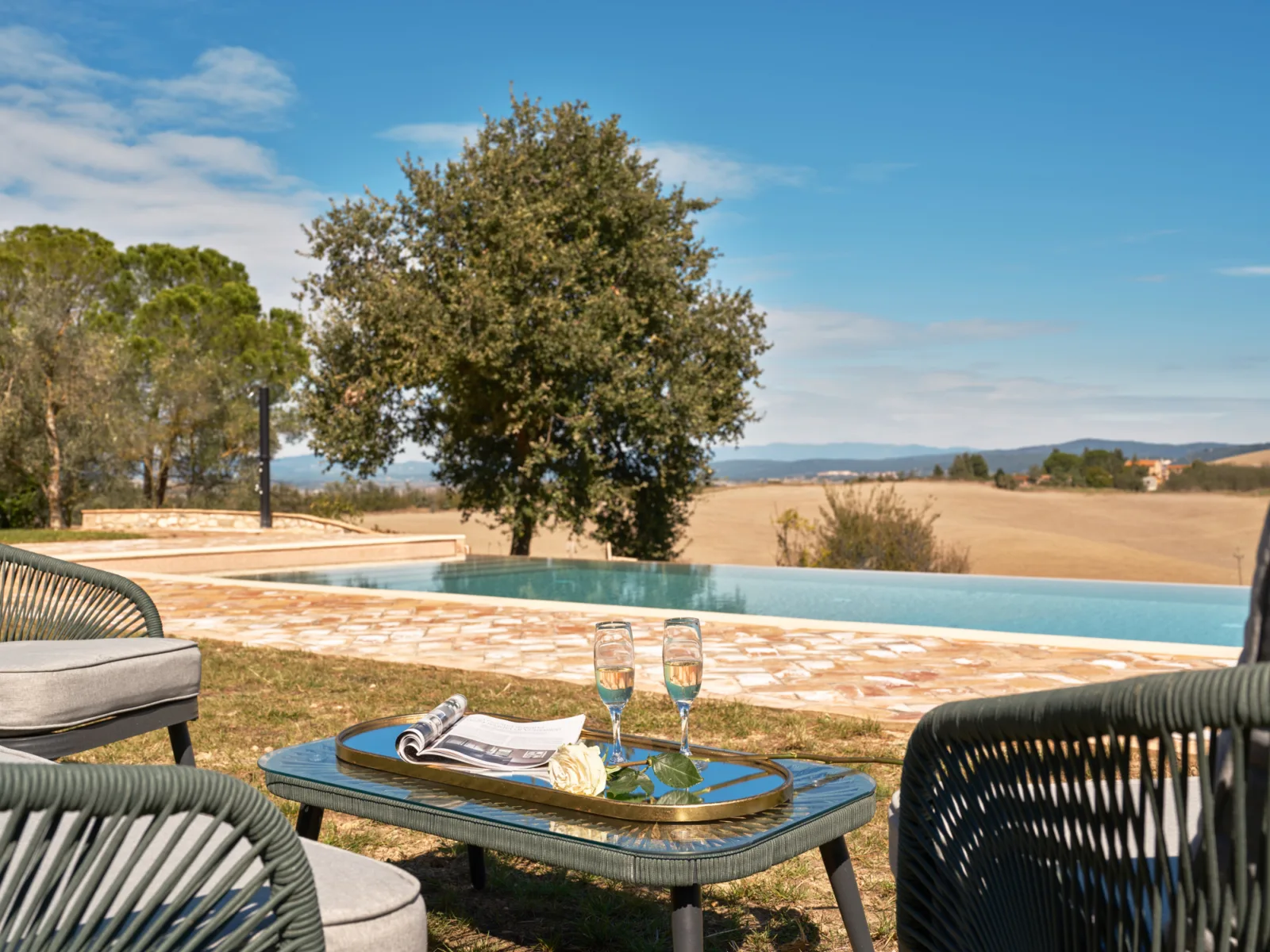 The infinity pool at Villa Eos, with a tray of champagne flutes and a magazine on a small green table, opening onto the Crete Senesi hills.